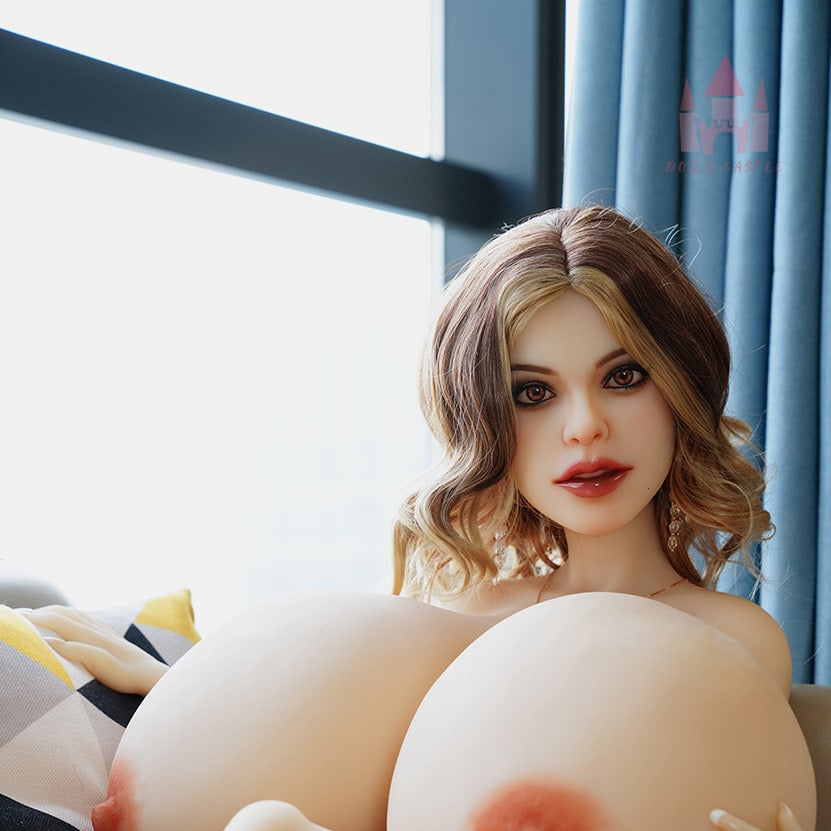A realistic mini sex doll with large breasts, brown hair, and white skin, seated and looking towards the camera.